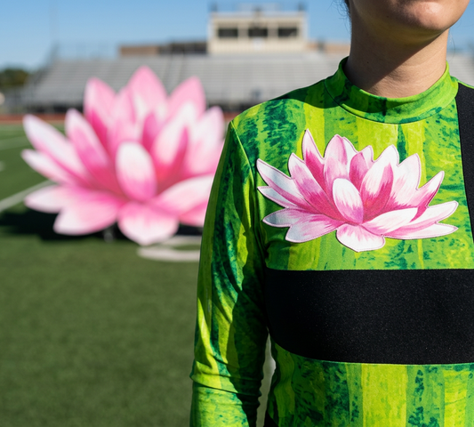 Close-up of a green and yellow watercolor-print marching band performance top featuring a pink lotus chest design and black waist band, displayed on a football field with a blurred stadium and matching lotus prop.